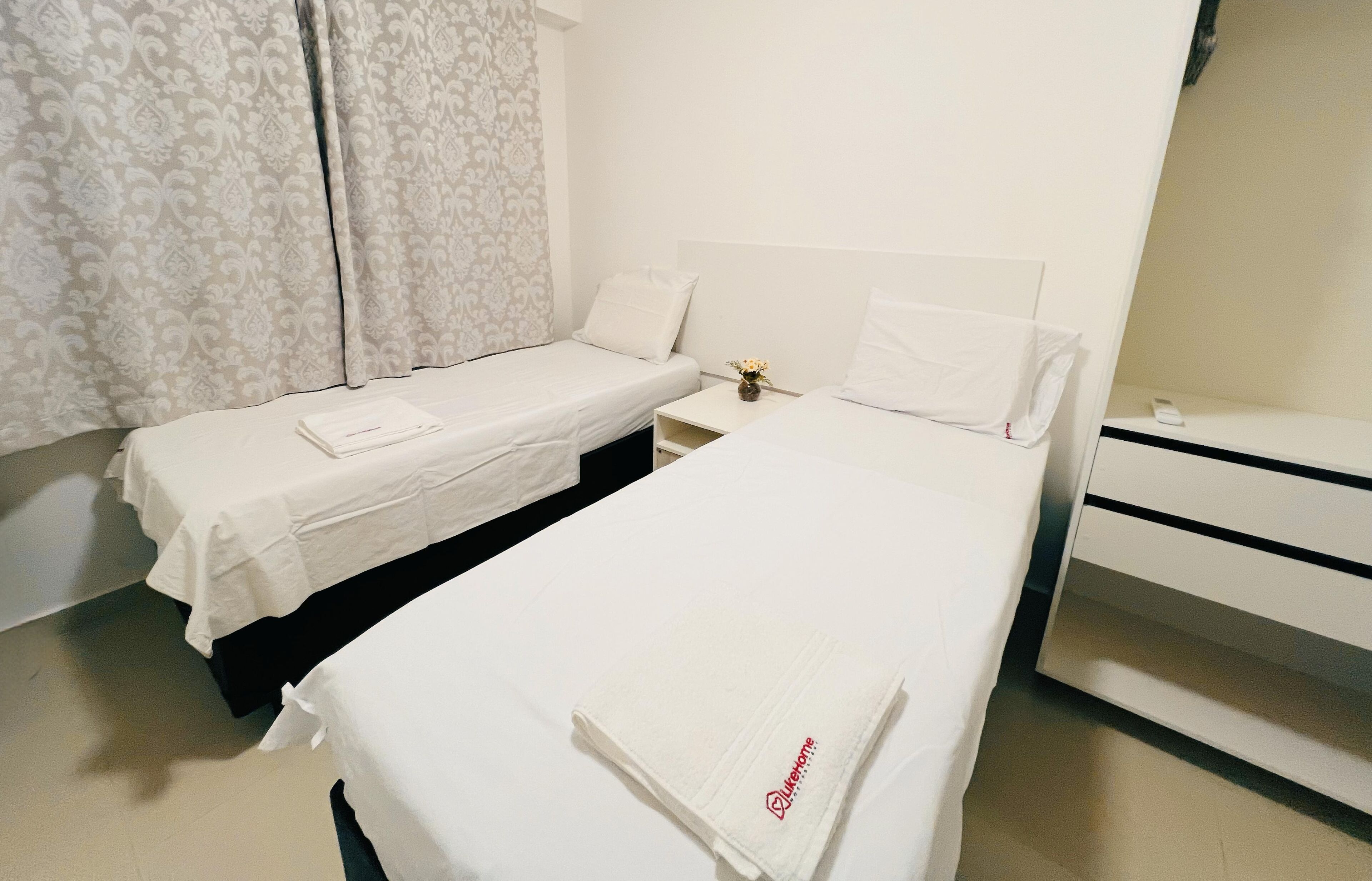 Una camera d'albergo con due letti singoli, un comodino con un vaso di fiori e una cassettiera bianca.