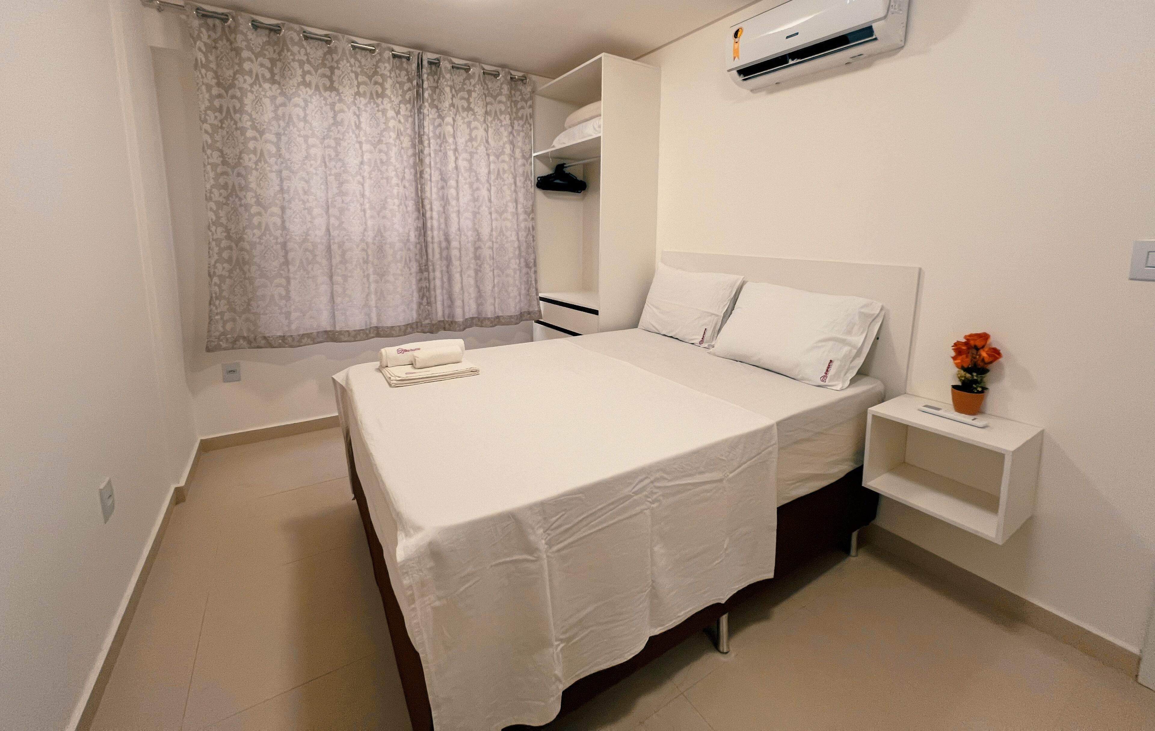 A neatly made bed with a white bedspread and pillows, a bedside table with a remote control, and a small potted plant.