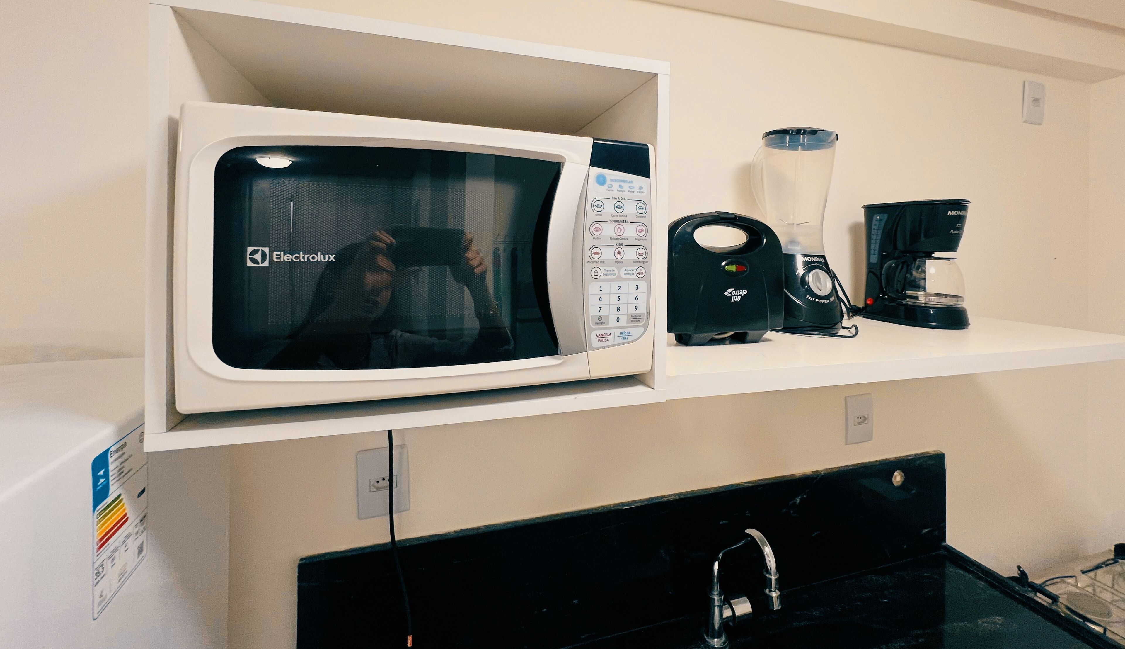 A microwave oven with a digital display and control panel, a blender, and a coffee maker on a white shelf.