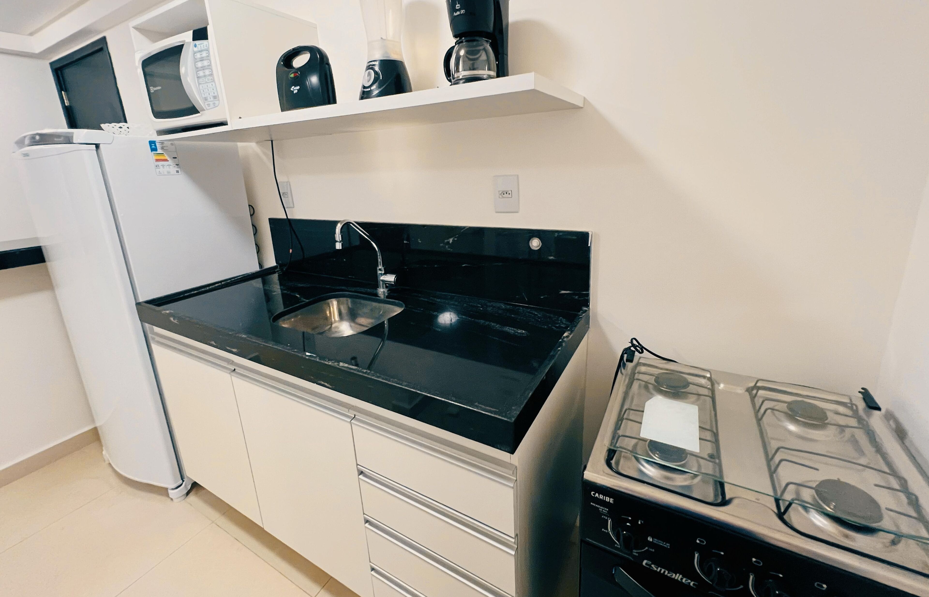 A compact kitchen with a black countertop, a stainless steel sink, a white refrigerator, a microwave, a coffee maker, and a gas stove.