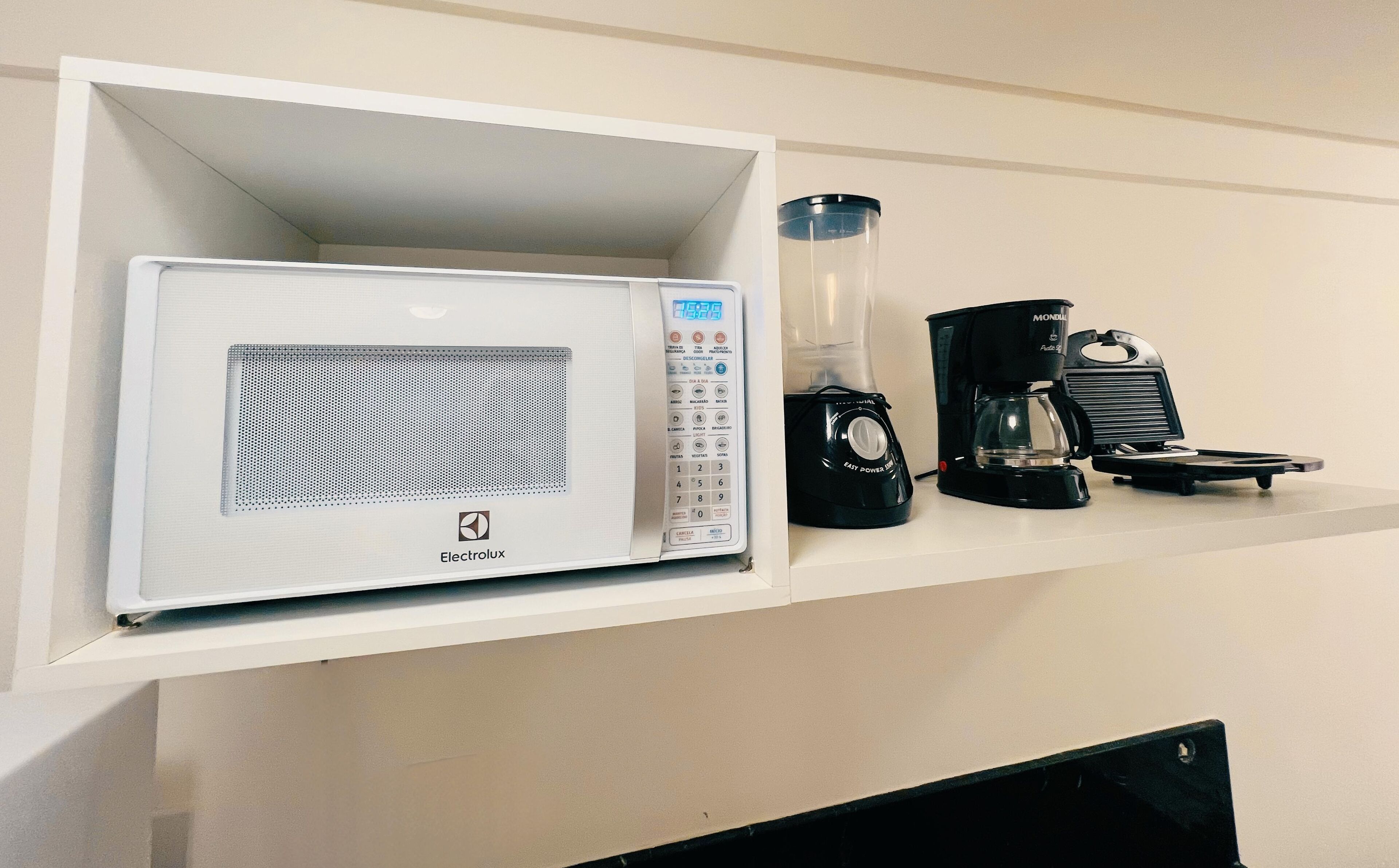 A microwave with a digital display, a coffee maker, and a blender on a shelf.