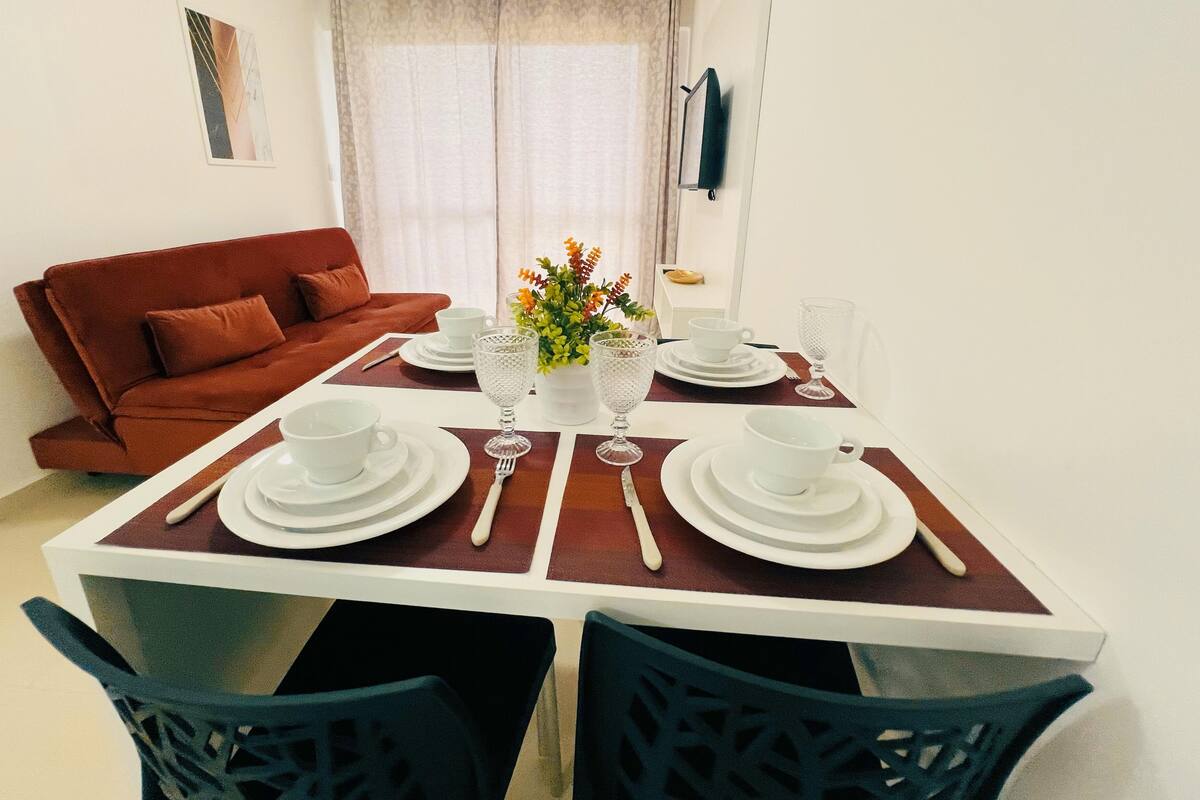 Ein Essbereich mit einem Tisch, gedeckt für vier Personen, inklusive weißem Geschirr, Besteck und Gläsern. Im Hintergrund befindet sich eine rote Couch und ein Fenster mit Vorhängen.