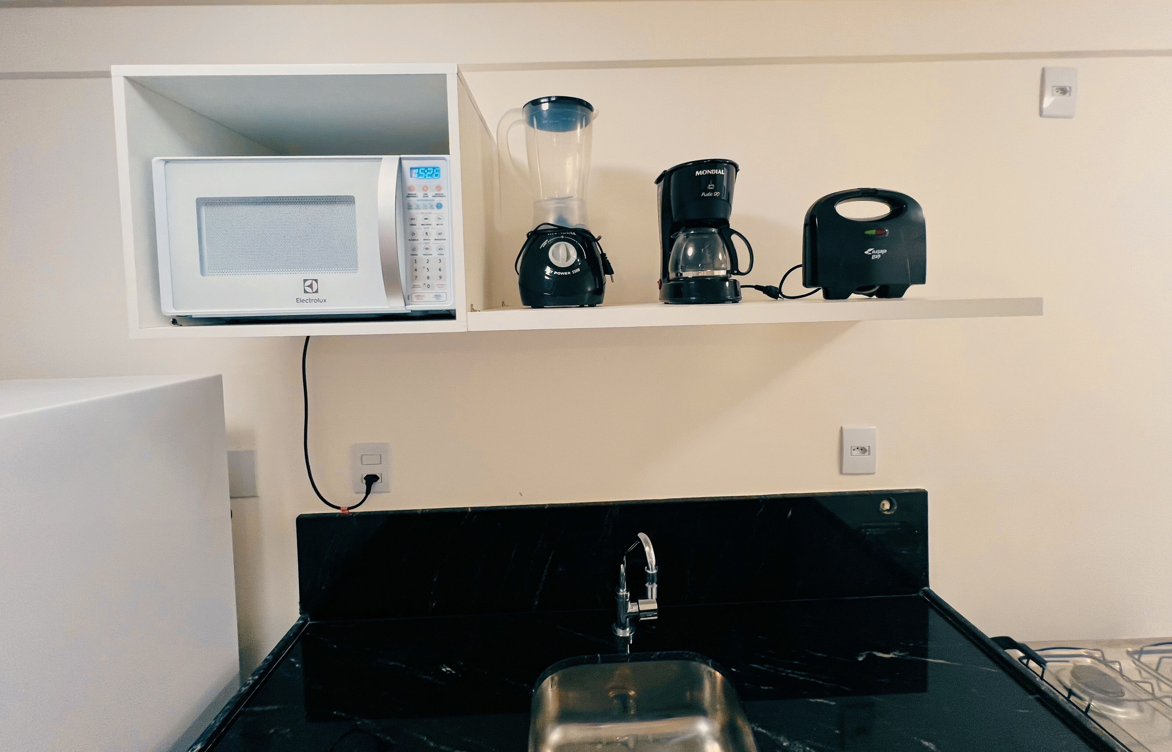 A kitchen with a microwave, blender, and coffee maker on a shelf, a black countertop, and a stainless steel sink.
