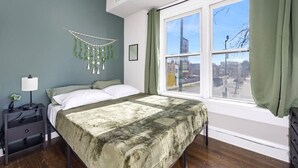 Une chambre à coucher avec un lit, une table de chevet, une lampe, une plante suspendue et une fenêtre avec des rideaux.