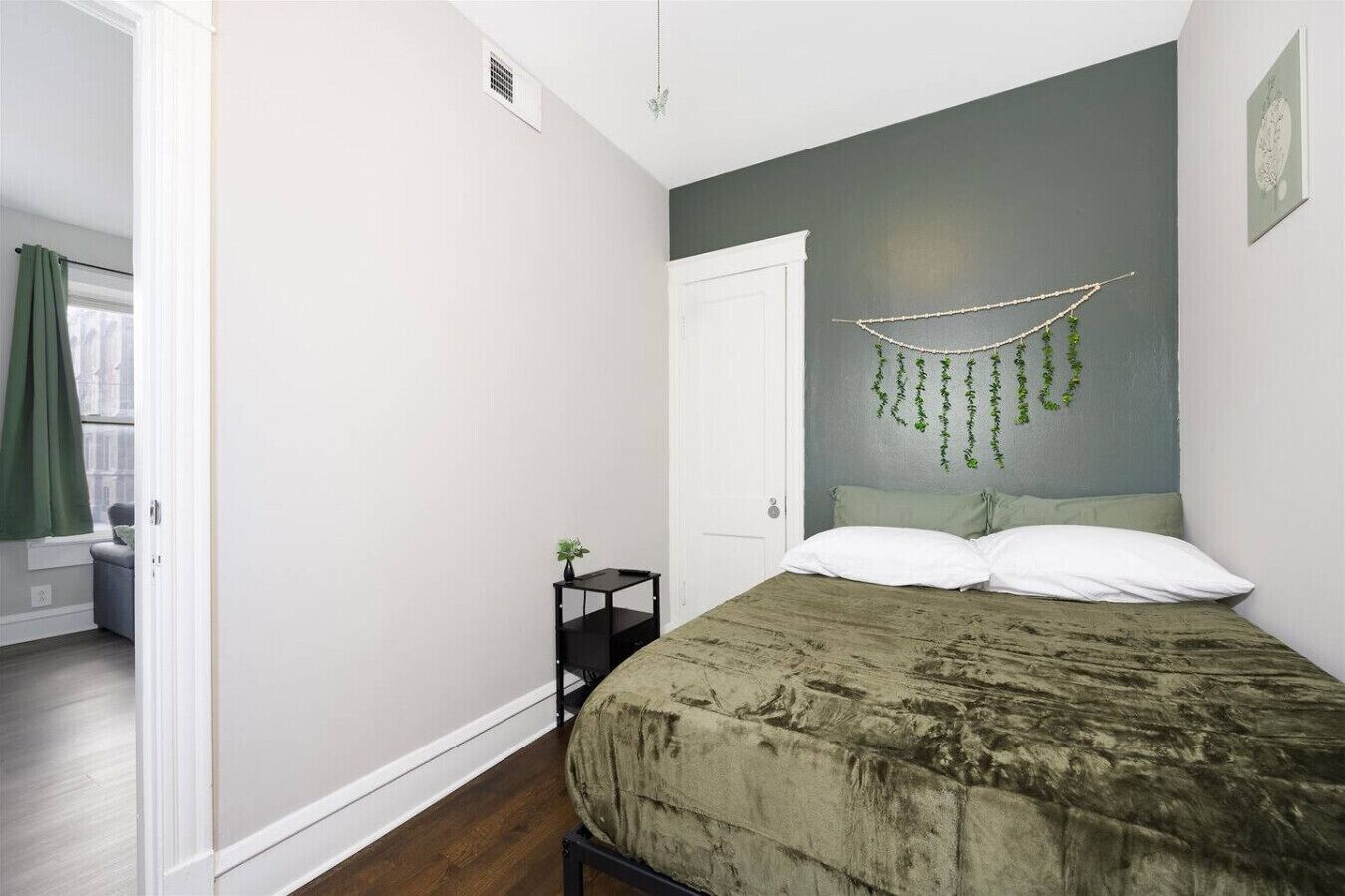 A bedroom with a bed, a bedside table, a hanging plant, and a window with curtains.