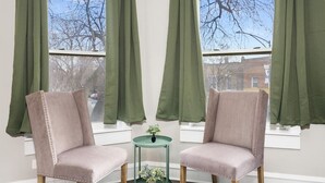 Deux chaises placées face à face, avec une petite table entre elles, une fenêtre avec des rideaux verts et une vue sur les arbres et les bâtiments à l’extérieur.