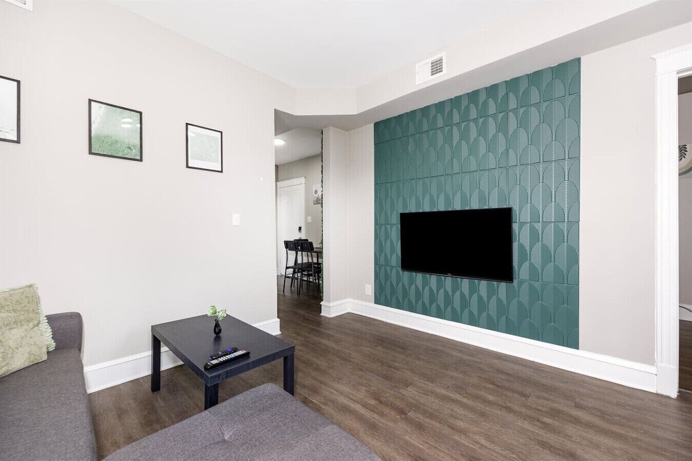 A living room with a flat-screen TV mounted on a teal-patterned wall, a dark coffee table, a gray sofa, and a dining area in the background.