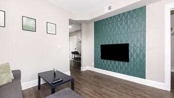 A living room with a flat-screen TV mounted on a teal-patterned wall, a dark coffee table, a gray sofa, and a dining area in the background.
