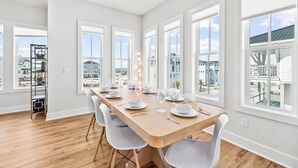 A spacious dining area with a long wooden table set for six, large windows offering views of the neighborhood, and a built-in shelf with decorative items. - The Sea Star: Sugar Sands Gated Community, Private Pool (Mexico Beach)