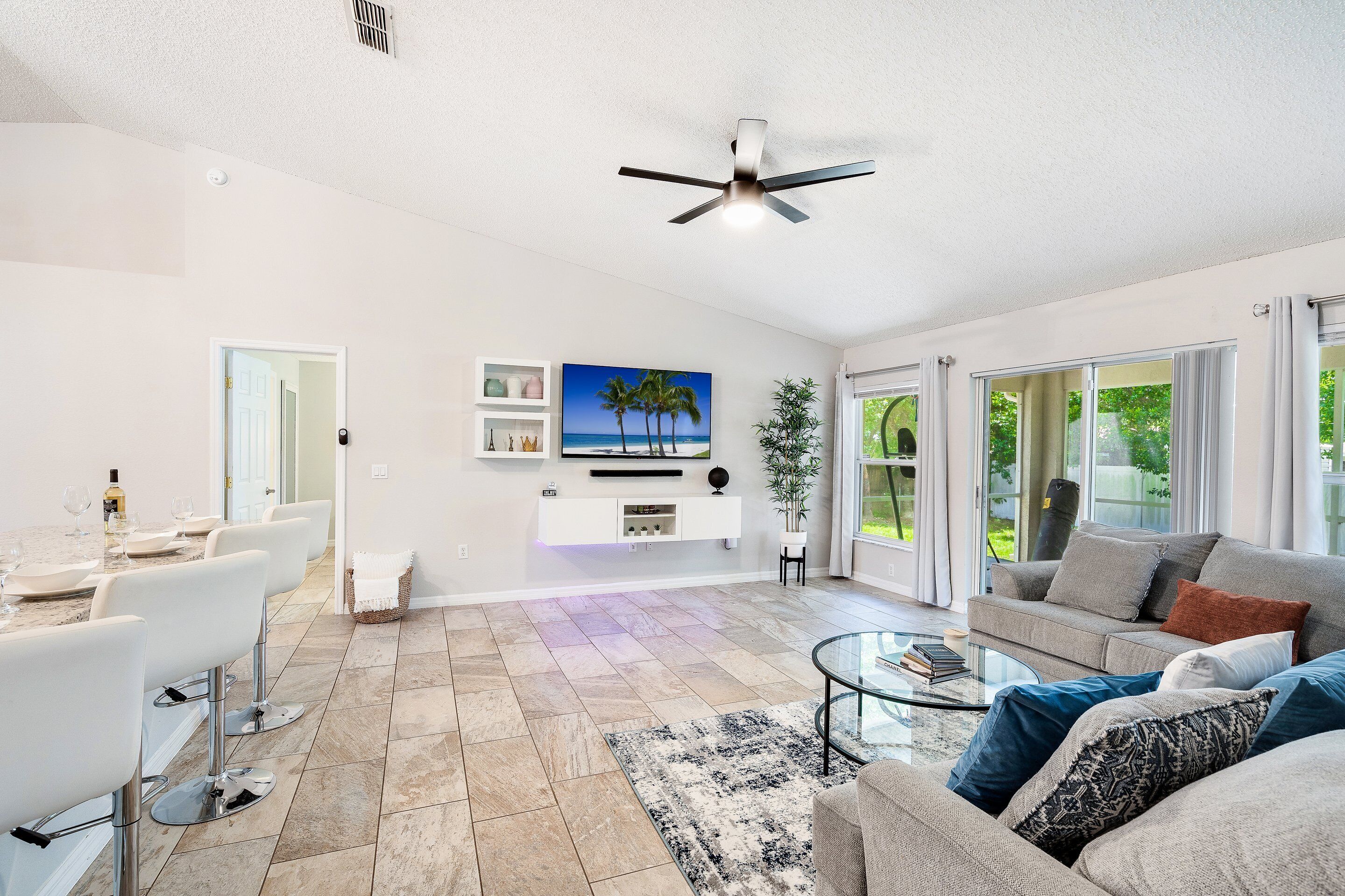 A spacious living room with a dining area, a ceiling fan, a flat-screen TV, and a large window offering a view of the outdoors.