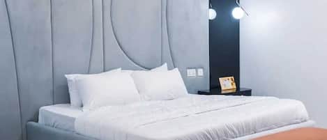Egy modern hálószoba, szürke, kárpitozott ágyvégfalával, fehér ágyával, melyen több párna található, barna padjával és egy lámpával felszerelt éjjeliszemlőjével.