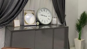 Een kamer met grijze gordijnen, een grijze kast met klokken en een potplant.