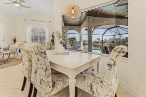 Ein Essbereich mit weißem Tisch und Stühlen, einem Deckenventilator und Blick auf einen Pool und Palmen.