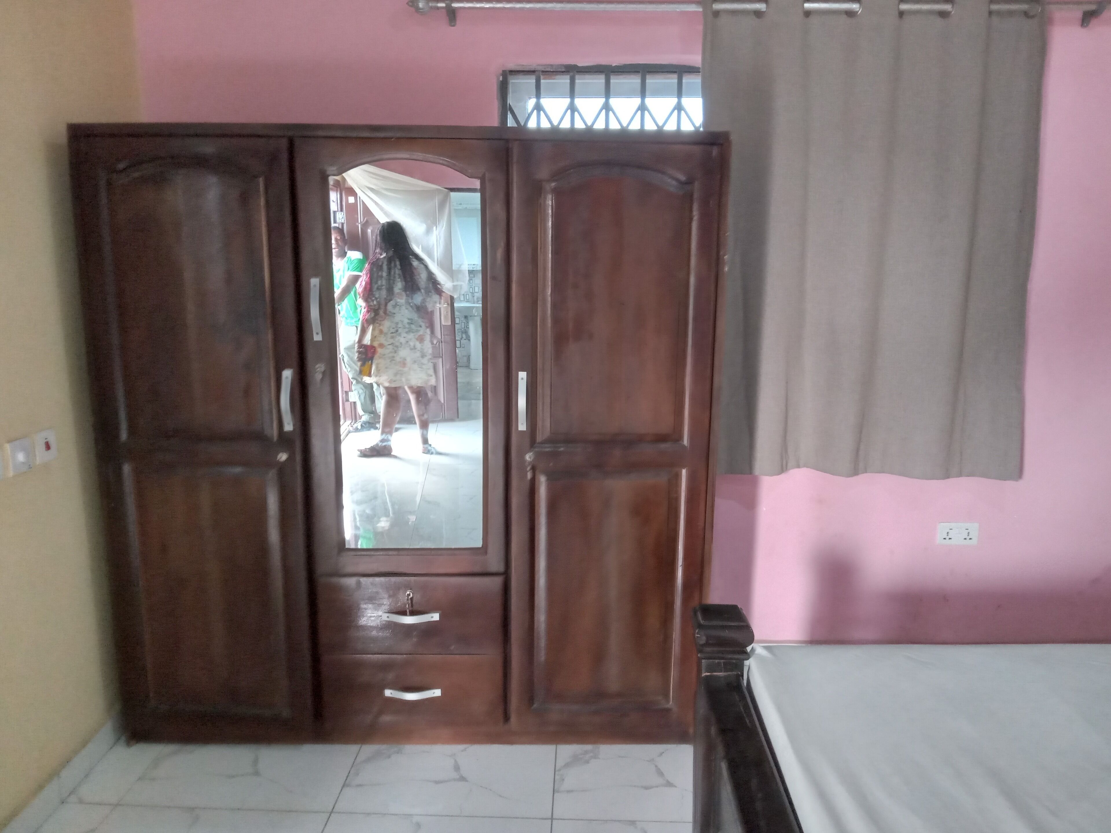 A room with a wooden wardrobe, a bed, and a pink wall.