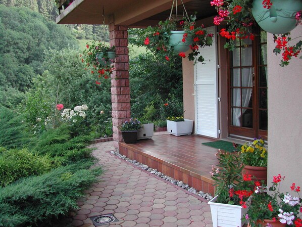 A covered porch with potted plants and flowers, a brick column, and a window with a white shutter.