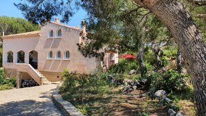 Traditional Mediterranean-style villa with a prominent staircase and arched windows, set amidst lush greenery and a stone pathway.