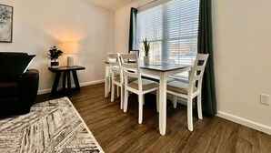 Ein Essbereich mit Tisch und Stühlen, ein kleiner Beistelltisch mit Lampe, ein dunkles Sessel und ein Fenster mit Vorhängen.