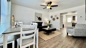 A modern living room with a dining area, a black sofa, a patterned armchair, a ceiling fan, and a dining table with chairs.