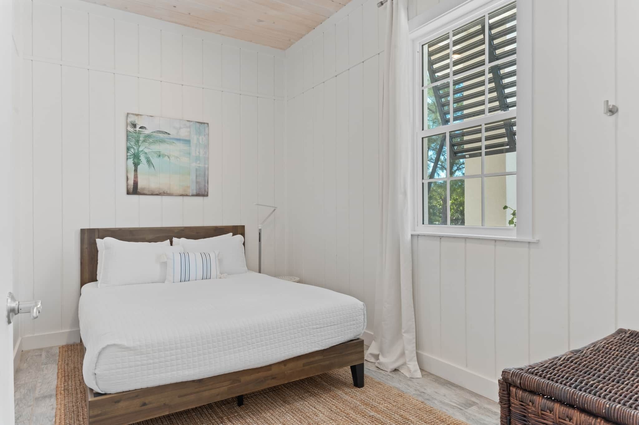 A bedroom with a wooden bed, a window with blinds, a painting on the wall, and a wicker chair.