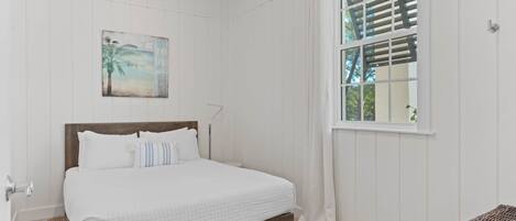 A bedroom with a wooden bed, a window with blinds, a painting on the wall, and a wicker chair.