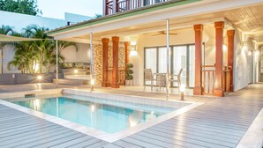 A swimming pool with a wooden deck, a covered patio area with seating, and a view of the surrounding greenery.