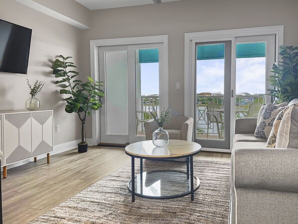 Un salon moderne avec une table basse ronde en verre, un canapé et un téléviseur fixé au mur. Deux portes-fenêtres coulissantes donnent sur un espace extérieur aménagé avec des chaises et une table.