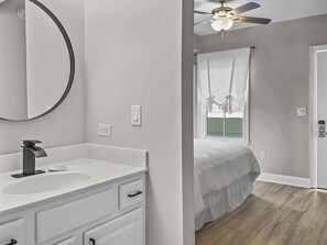 Une chambre à coucher avec un lit, une fenêtre avec des rideaux, un ventilateur de plafond et une salle de bain avec un lavabo et un miroir.