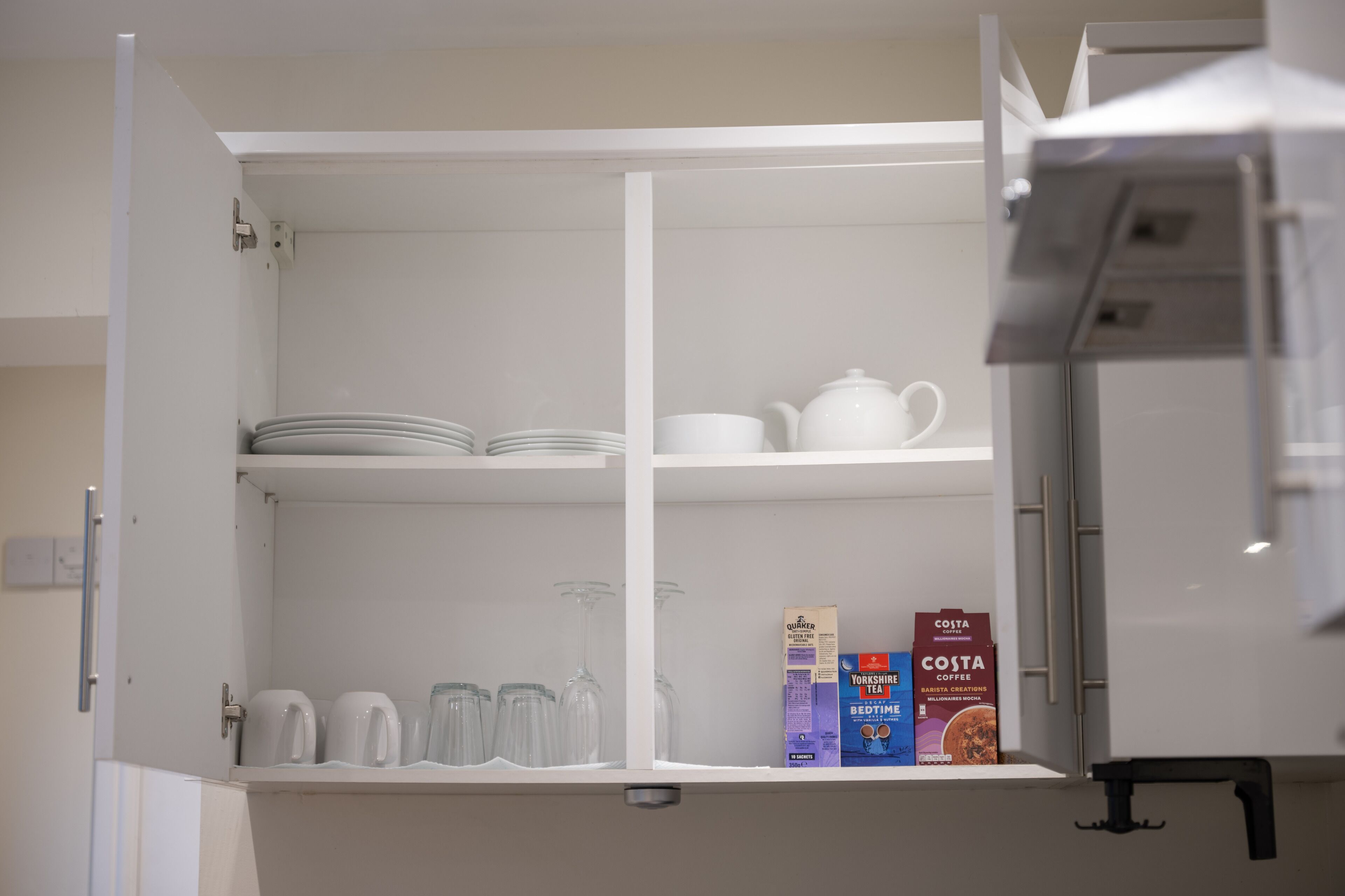 A kitchen with open cabinets displaying tea and coffee boxes, glassware, and plates.