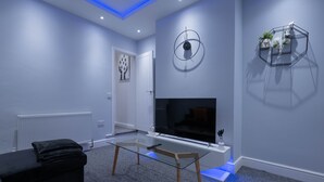 Un salon moderne avec une table basse en verre, un canapé noir, un téléviseur fixé au mur et une étagère murale aux motifs géométriques.