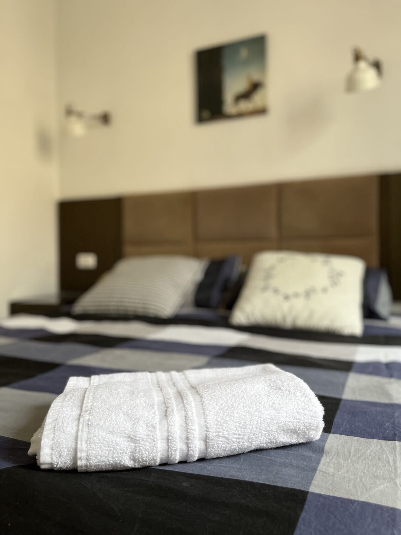 Una cama bien hecha con colcha a cuadros, una toalla blanca doblada y un cabecero con un cuadro enmarcado encima.