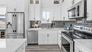 A modern kitchen with white cabinetry, stainless steel appliances, and a black tiled backsplash. - Cold Pass Cottage - 3BR, panoramic views (Freeport)