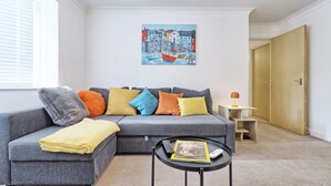 A modern living room with a grey sectional sofa, colorful cushions, a small round coffee table with a magazine, and a painting of a harbor scene on the wall.