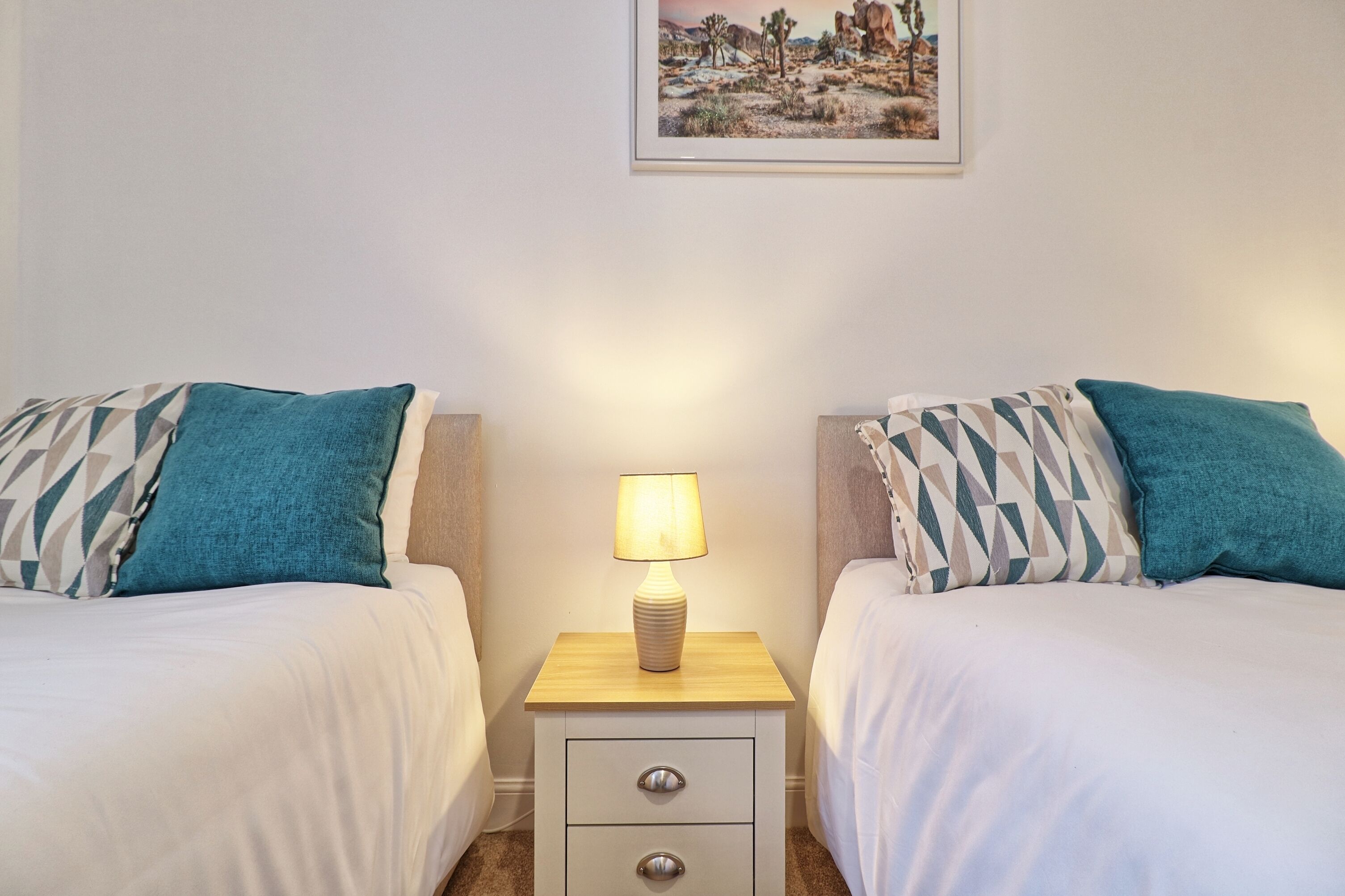 Een hotelkamer met twee bedden, elk voorzien van geborduurde kussens, een nachtkastje met een lamp en een lijst met een afbeelding aan de muur.