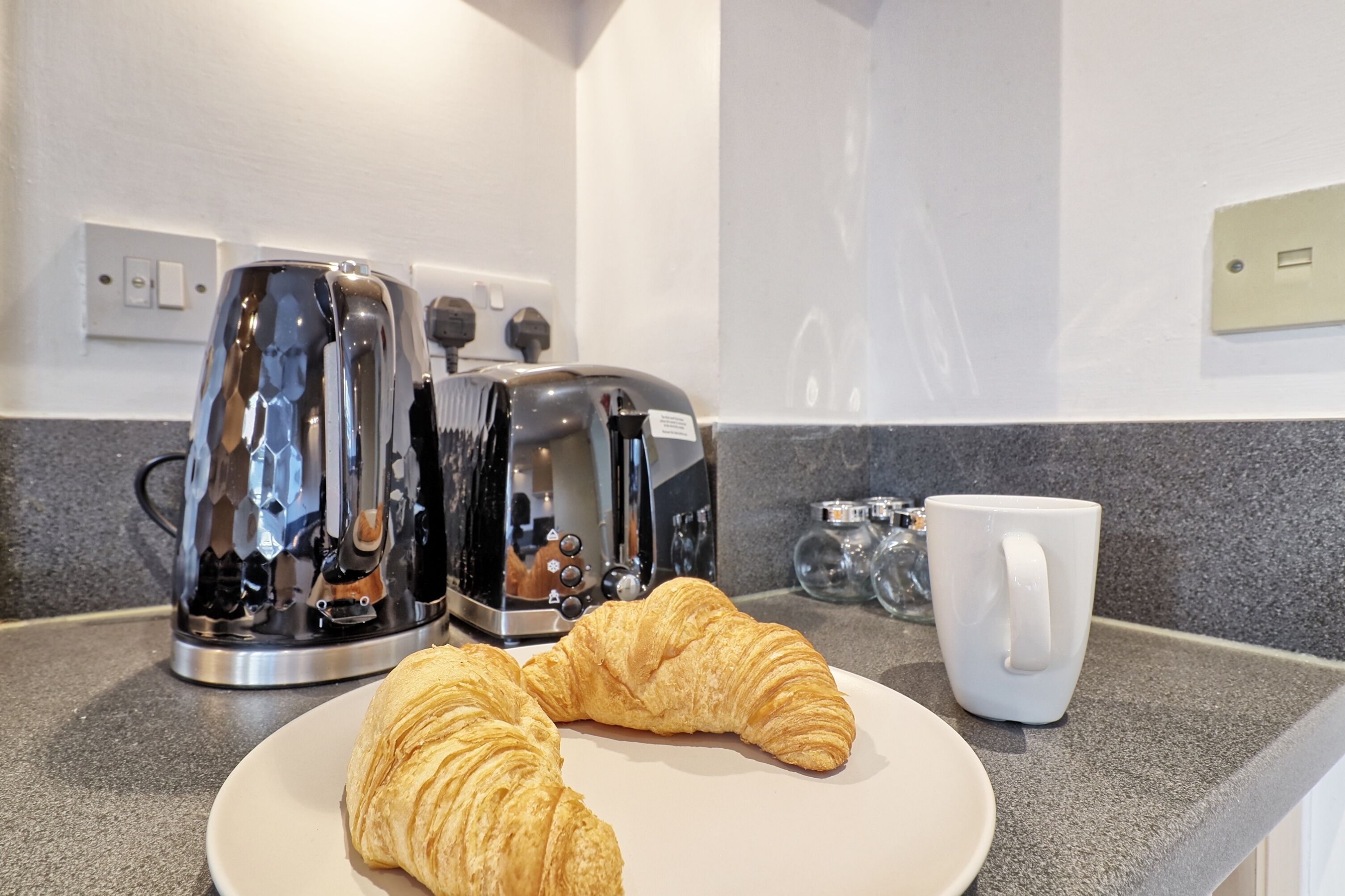 A kitchen counter with a plate of croissants, a coffee maker, and a white mug.
