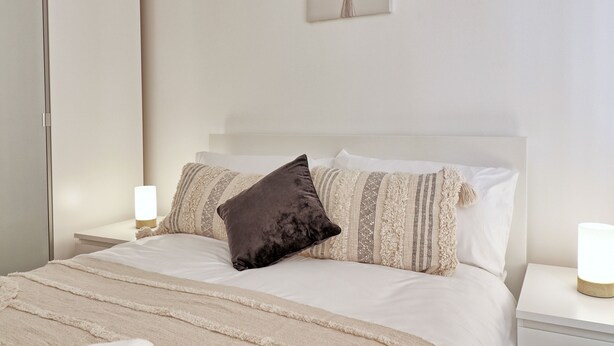 A neatly made bed with a dark pillow, white bedding, and a bedside lamp on a white nightstand.