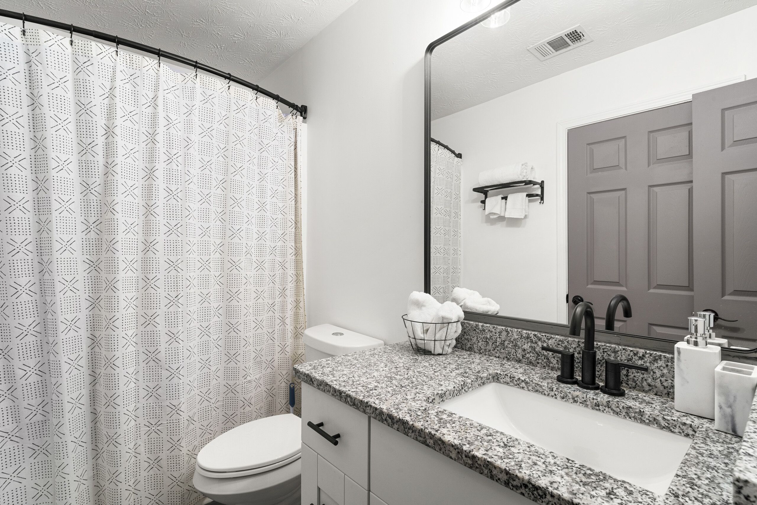 A bathroom with a white toilet, a granite sink, a black faucet, a towel rack, and a shower curtain with a geometric pattern.