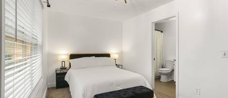 A bedroom with a bed, bedside tables, a bench, a ceiling fan, and a bathroom visible through an open door.