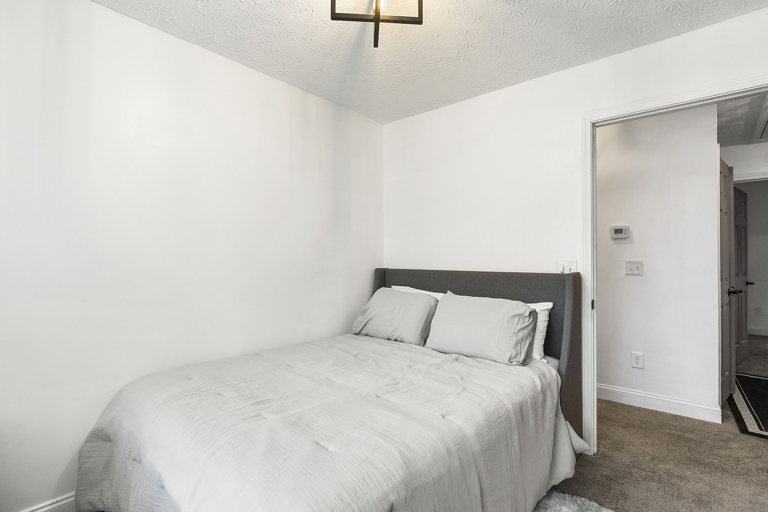 A bedroom with a bed, white walls, a ceiling light fixture, and a door leading to another room.