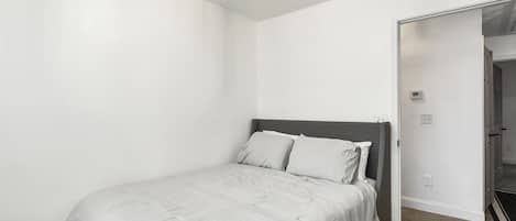 A bedroom with a bed, white walls, a ceiling light fixture, and a door leading to another room.