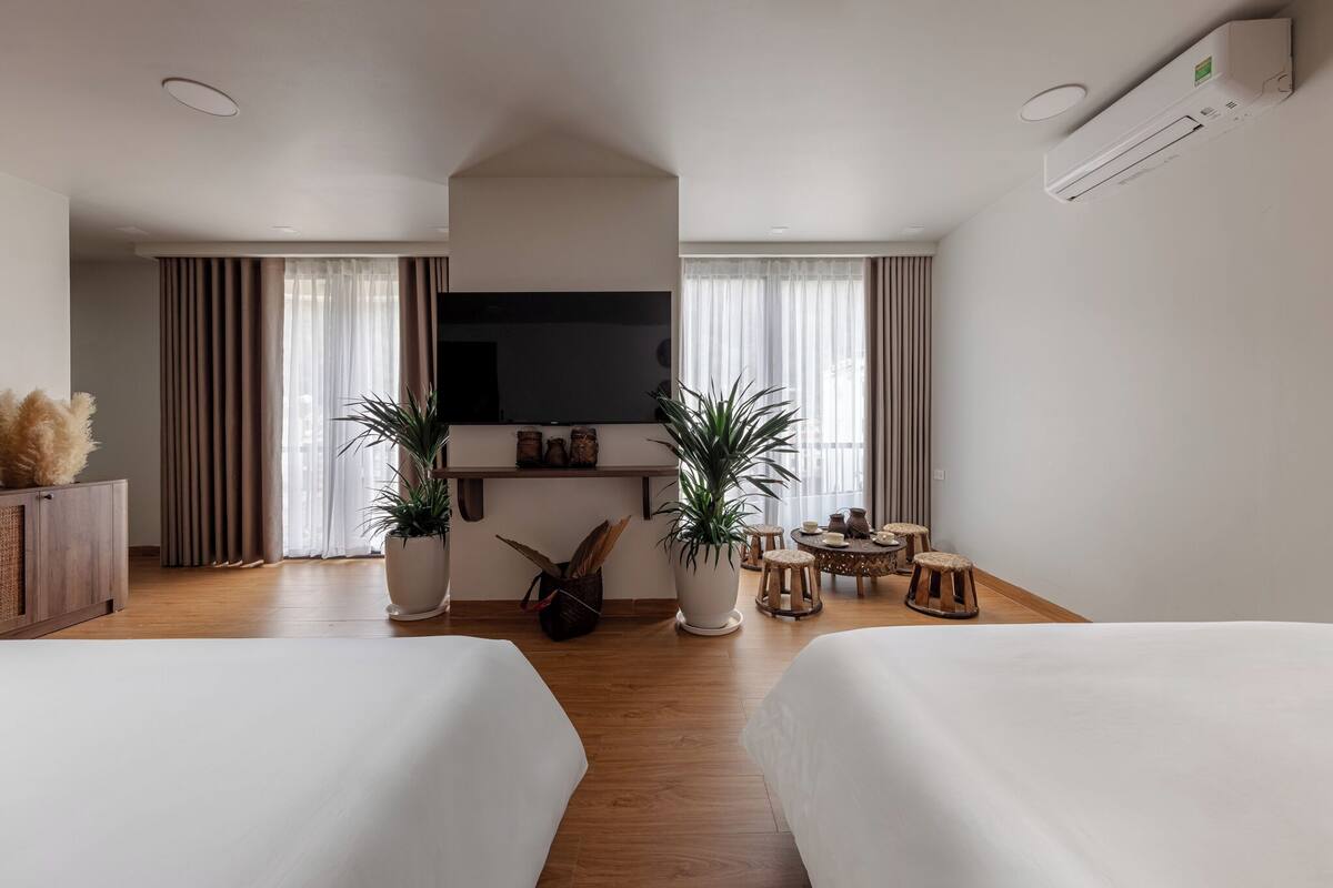 A hotel room with two beds, a flat-screen TV, a potted plant, a small round table with stools, and a wooden cabinet.