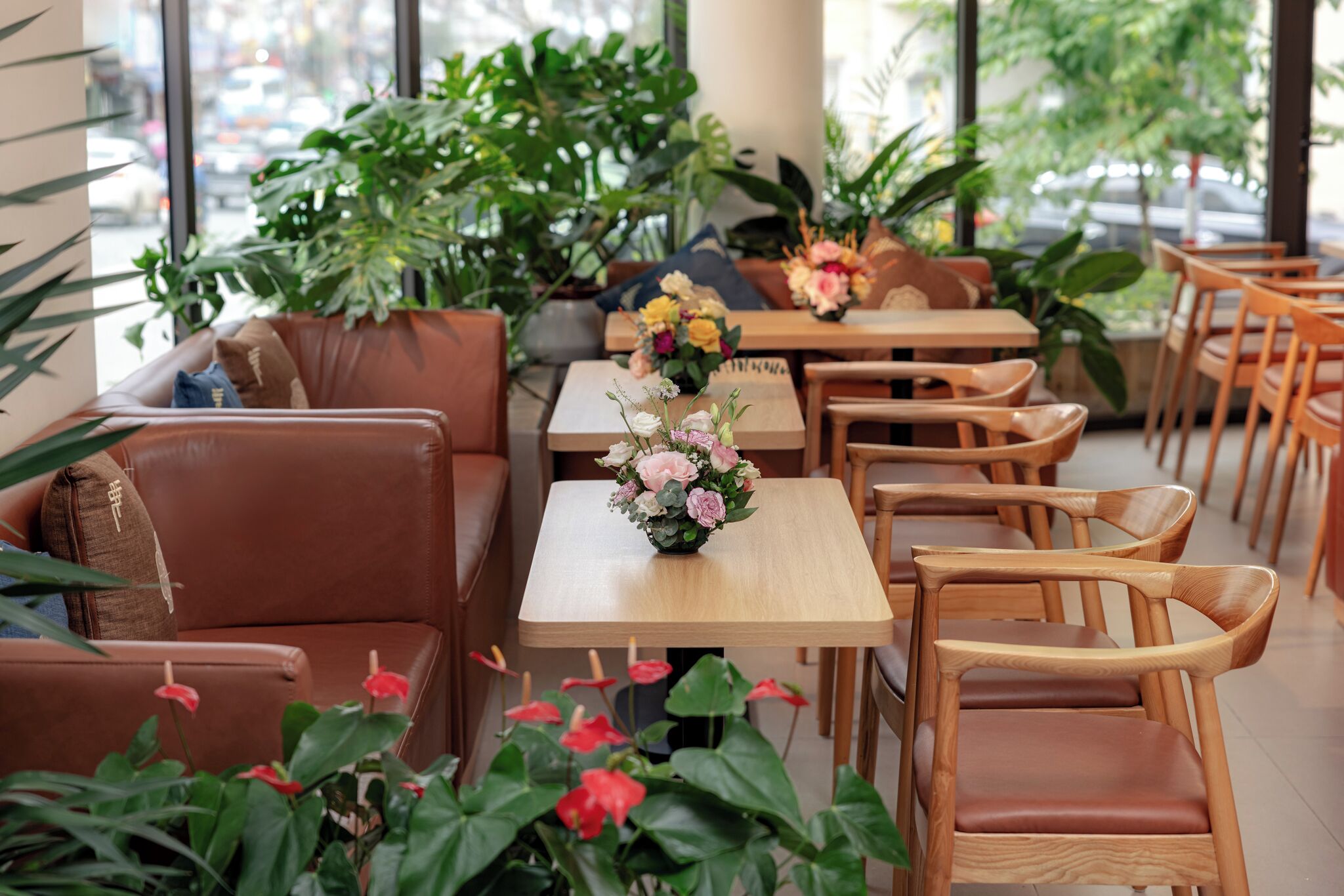 Een café met houten tafels en stoelen, planten in potten en uitzicht op de straat door grote ramen.