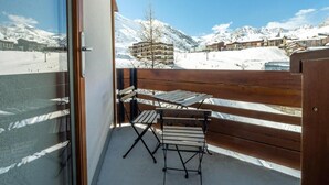 A balcony with a wooden bench and table, offering a view of snow-covered mountains and buildings.