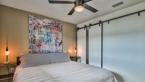 Un dormitorio con una cama, un ventilador de techo, una lámpara de pared y un cuadro abstracto grande.