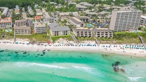 Vista aérea de un complejo turístico costero con varios edificios, una playa de arena y aguas turquesas cristalinas.