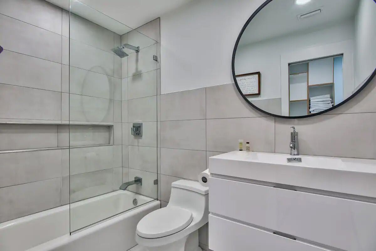 Une salle de bain moderne avec une douche à parois de verre, des toilettes blanches et un miroir rond placé au-dessus d’un meuble-lavabo blanc.