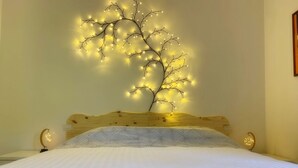 Una cama con edredón blanco y almohada estampada, cabecera de madera y lámpara de pared con forma de árbol iluminado.
