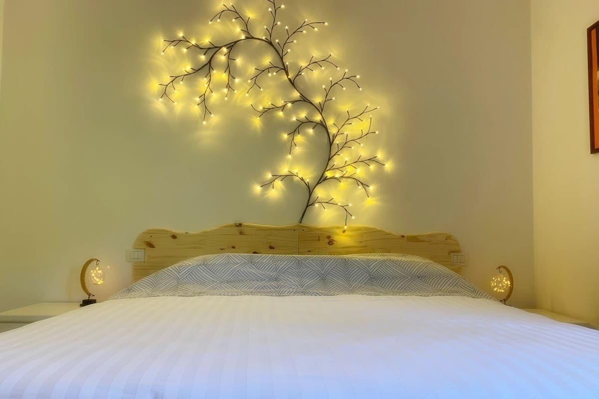 Uma cama com edredom branco e almofada estampada, cabeceira de madeira e luminária fixada na parede que lembra uma árvore com luzes.
