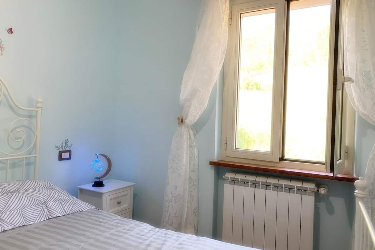 Quarto com cama, mesa de cabeceira, janela com cortinas e radiador.