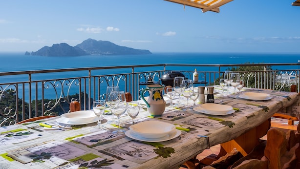 Mesa de comedor preparada con platos, vasos y una jofaina, con vistas a un paisaje costero.