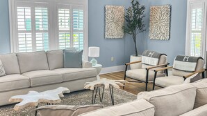 A living room with a beige sectional sofa, two armchairs, a coffee table, and a floor lamp. There are two large windows with blinds, a ceiling fan, and decorative wall art. - Remodeled 5 bedroom house with bikes – walking distance to the beach  (Santa Rosa Beach)